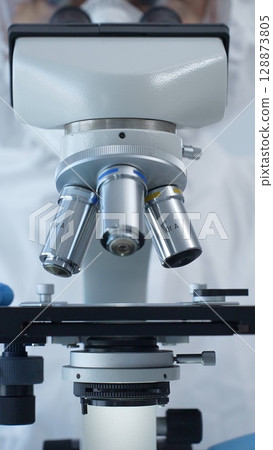 Close up of medical virologist in a hazmat suit, mask, and gloves, diligently working at a microscope in a laboratory researching viruses. Medicine and science concept Close up of medical virologist in a hazmat suit, mask, and gloves, diligently working at a microscope in a laboratory researching viruses. Medicine and science concept 128873805