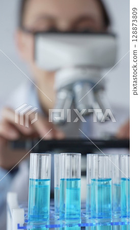 Close up of test tubes with blue liquid while scientist focusing on analyzing samples under a microscope in a laboratory setting. Medicine, healthcare and science concept 128873809