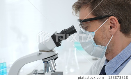 Scientist wearing mask and gloves using microscope doing scientific research in a laboratory with test tubes containing blue liquid. Medicine, healthcare and science concept 128873818
