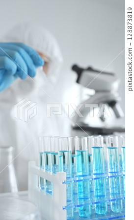 Scientific researcher wearing protective equipment, carefully pipetting liquid into test tubes while conducting medical research experiment. Medicine, healthcare and science concept Scientific researcher wearing protective equipment, carefully pipetting liquid into test tubes while conducting medical research experiment. Medicine, healthcare and science concept 128873819