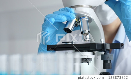 Close up of scientist's gloved hands adjusting microscope, conducting research in laboratory with test tubes containing blue liquid in background. Medicine, healthcare and science concept 128873832