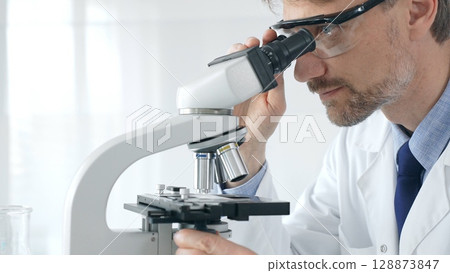 Adult male scientific researcher analyzing sample through microscope, wearing protective gear in high tech laboratory environment. Healthcare, medicine and science concept 128873847