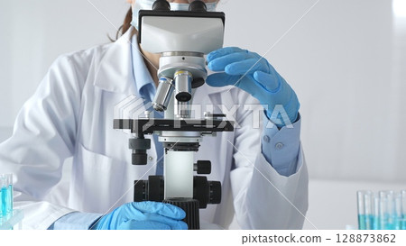 A female scientist in protective gear adjusts a microscope lens and accurately analyzes medical research samples in a sterile laboratory setting A female scientist in protective gear adjusts a microscope lens and accurately analyzes medical research samples in a sterile laboratory setting 128873862