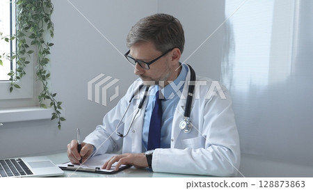 Doctor man documenting patient information, writing notes on clipboard while seated at desk, laptop and window side greenery framing clinical workspace. Medicine and health care Doctor man documenting patient information, writing notes on clipboard while seated at desk, laptop and window side greenery framing clinical workspace. Medicine and health care 128873863