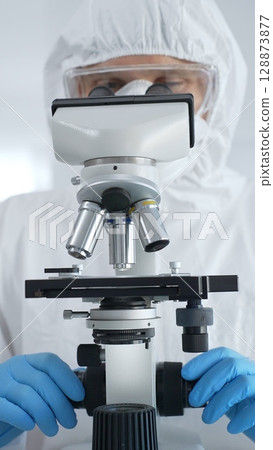 Male virologist in a hazmat suit, mask, and blue gloves, diligently working at a microscope in a laboratory researching viruses. Medicine, science concept Male virologist in a hazmat suit, mask, and blue gloves, diligently working at a microscope in a laboratory researching viruses. Medicine, science concept 128873877