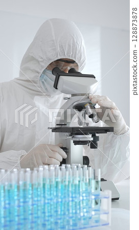 A medical virologist in protective gear examines viral samples under a microscope while conducting research in a laboratory setting A medical virologist in protective gear examines viral samples under a microscope while conducting research in a laboratory setting 128873878