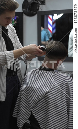 Professional barber carefully trimming customer's hair with electric clipper, providing a stylish and precise haircut in a modern barbershop environment Professional barber carefully trimming customer's hair with electric clipper, providing a stylish and precise haircut in a modern barbershop environment 128873879