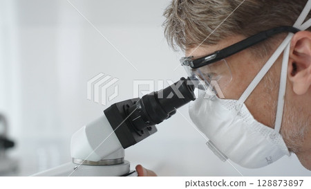 Male scientific researcher wearing protective gear examining sample through microscope, performing detailed laboratory investigation. Medicine and science concept Male scientific researcher wearing protective gear examining sample through microscope, performing detailed laboratory investigation. Medicine and science concept 128873897