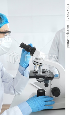 Two scientists in lab coats, face masks, and protective glasses conducting research with a microscope in a laboratory. Medicine and science concept Two scientists in lab coats, face masks, and protective glasses conducting research with a microscope in a laboratory. Medicine and science concept 128873904