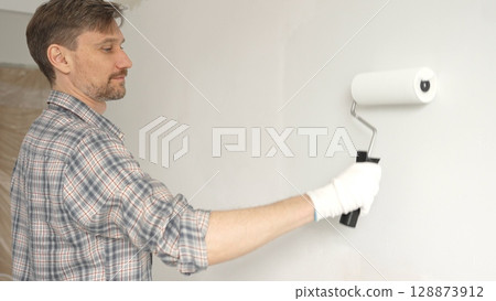 Man professional painter wearing checkered shirt, is rolling white paint on apartment wall with paint roller, transforming interior space with steady strokes during comprehensive home renovation Man professional painter wearing checkered shirt, is rolling white paint on apartment wall with paint roller, transforming interior space with steady strokes during comprehensive home renovation 128873912
