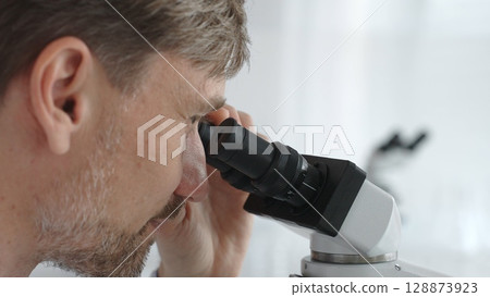 Close up of a man scientist carefully observing through a microscope in a brightly lit laboratory, conducting research for developing new medicines and treatments. Medicine and science concept 128873923