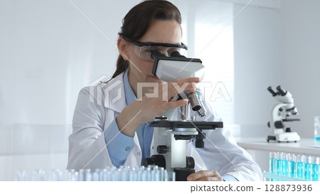 Female scientist wearing lab coat and protective glasses using microscope, analyzing samples in test tubes in modern laboratory, conducting scientific research 128873936