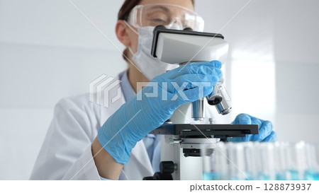 Woman scientist wearing protective gear is checking microscope, analyzing blue test tubes in modern laboratory research setting. Medicine, healthcare and science concept 128873937