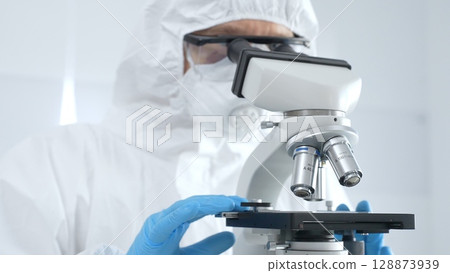 Medical virologist in a hazmat suit, mask, and blue protective gloves, carefully working at a microscope in a laboratory researching viruses. Healthcare and science concept Medical virologist in a hazmat suit, mask, and blue protective gloves, carefully working at a microscope in a laboratory researching viruses. Healthcare and science concept 128873939