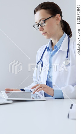 Middle aged doctor woman wearing lab coat and stethoscope using digital tablet with laptop and medical records on desk in medical office, showcasing modern healthcare technology. Medicine and health 128873943