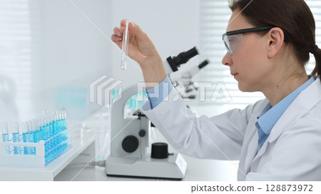 Female researcher examining clear liquid in test tube, sitting near microscopes and lab equipment within sterile scientific workspace. Medicine, healthcare and science concept 128873972