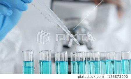 Scientific glassware containing blue solution, researcher carefully dispensing liquid via pipette near microscope within sterile laboratory setting, close up. Medicine, healthcare and science concept Scientific glassware containing blue solution, researcher carefully dispensing liquid via pipette near microscope within sterile laboratory setting, close up. Medicine, healthcare and science concept 128873990