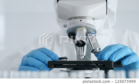 Scientific researcher or virologist wearing protective gear is tuning microscope lenses opposite the lab tubes with blue liquid in laboratory setting. Medicine, healthcare and science concept 128873999