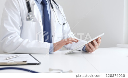 Doctor man wearing white medical coat and stethoscope using digital tablet while sitting at desk in medical office, with medical chart and pen in the foreground. Medicine concept 128874010