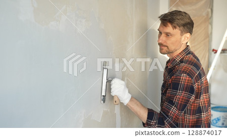 Male construction worker wearing protective gloves smoothing plaster onto wall surface during home renovation using professional trowel tool 128874017