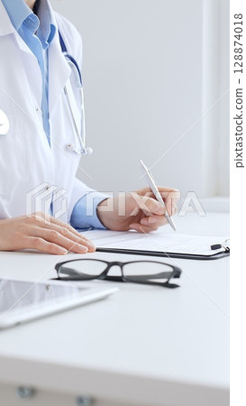 Unknown female doctor completing healthcare paperwork, clipboard positioned near laptop and stethoscope in well lit clinical workspace by window. Medicine and health care Unknown female doctor completing healthcare paperwork, clipboard positioned near laptop and stethoscope in well lit clinical workspace by window. Medicine and health care 128874018