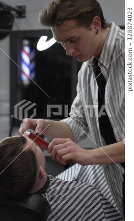 Professional barber trimming customer's hair with electric clipper, focusing on precise nape grooming within contemporary barbershop interior design Professional barber trimming customer's hair with electric clipper, focusing on precise nape grooming within contemporary barbershop interior design 128874023
