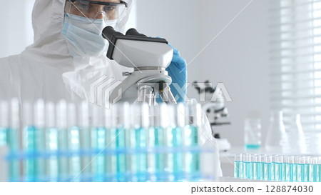 A male doctor in a protective suit and gloves uses a microscope while analyzing samples, conducting research in a laboratory. Close-up of test tubes with blue liquid 128874030