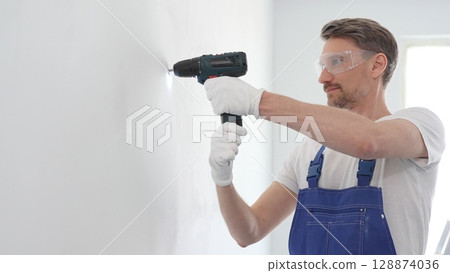 Professional handyman wearing blue overall, safety glasses and gloves, is using cordless drill to make hole in white wall during renovation or installation work 128874036