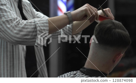 Skilled barber trimming customer's hair using professional scissors and comb, working in contemporary barbershop with vintage pole visible near workspace 128874098