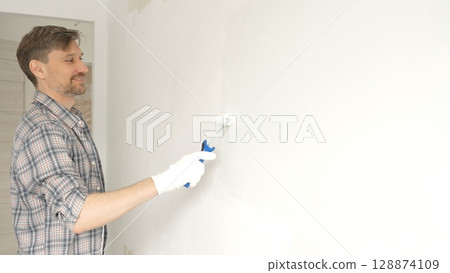 Professional male painter wearing checkered shirt, is rolling white paint on apartment wall, transforming interior space with steady strokes during comprehensive home renovation 128874109