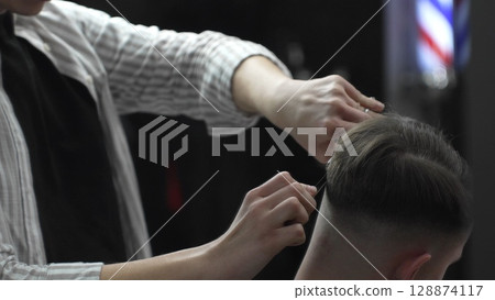 Skilled barber trimming customer's hair using professional scissors and comb, working in contemporary barbershop with vintage pole visible near workspace Skilled barber trimming customer's hair using professional scissors and comb, working in contemporary barbershop with vintage pole visible near workspace 128874117