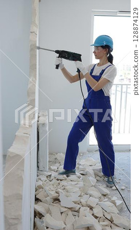 Female construction worker demolishing interior wall with rotary hammer, wearing safety glasses and hardhat, creating substantial concrete debris pile during demolition process 128874129