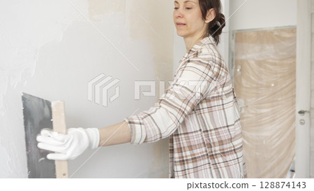 Woman applying plaster to wall with putty knife, wearing protective gloves and creating smooth surface during home renovation work 128874143