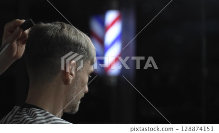 Professional barber grooming male client's hair with scissors, vintage barber pole framing traditional barbershop setting 128874151