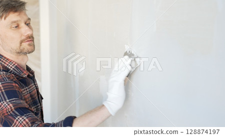 Male construction worker spreading plaster on white wall, wearing protective gloves during home renovation project with putty knife 128874197