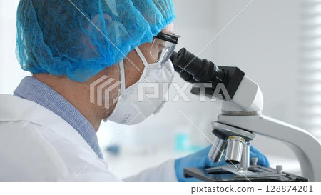 Male scientist wearing protective gear examining microscope slide, analyzing research sample in sterile laboratory environment. Health care and science concept Male scientist wearing protective gear examining microscope slide, analyzing research sample in sterile laboratory environment. Health care and science concept 128874201