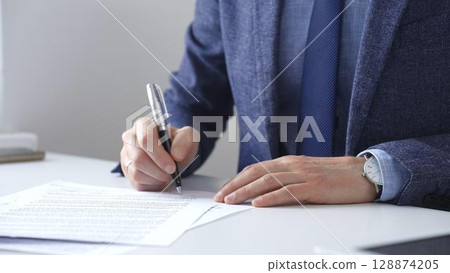 Professional businessman signing contract with silver pen, wearing tailored blue suit, business documents and digital tablet nearby, close up of hands. Business people concept Professional businessman signing contract with silver pen, wearing tailored blue suit, business documents and digital tablet nearby, close up of hands. Business people concept 128874205