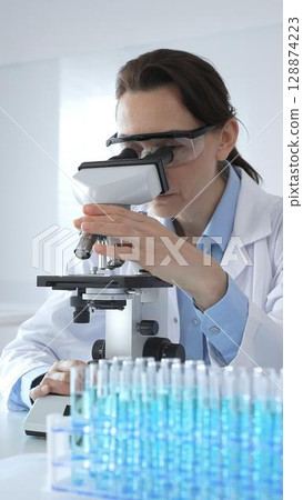 Female scientist examining microscope slide, wearing protective gear in laboratory, researching pharmaceutical breakthrough with blue tinted test tube specimens 128874223