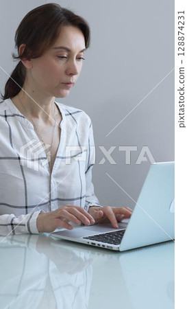 Professional businesswoman working on laptop, sitting at glass desk in bright contemporary workspace, embodying corporate success and concentration. Business people concept. 128874231
