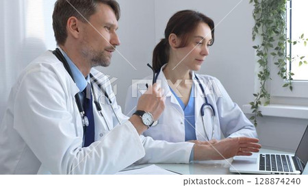 Two doctors are examining medical records on a laptop, engaging in a focused discussion about patient data within a hospital or clinical setting. Medicine and health care concept Two doctors are examining medical records on a laptop, engaging in a focused discussion about patient data within a hospital or clinical setting. Medicine and health care concept 128874240