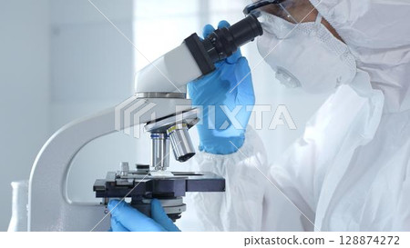 Medical virologist in a hazmat suit, mask, and gloves, diligently working at a microscope in a laboratory researching viruses. Healthcare and science concept 128874272