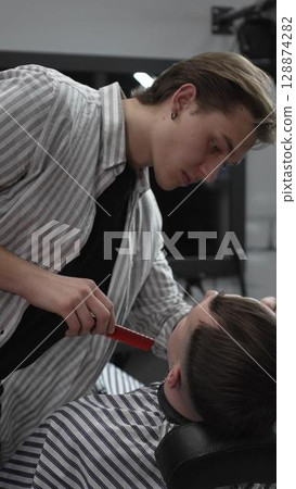 Professional barber trimming customer's hair with electric clipper, focusing on precise nape grooming within contemporary barbershop interior design Professional barber trimming customer's hair with electric clipper, focusing on precise nape grooming within contemporary barbershop interior design 128874282