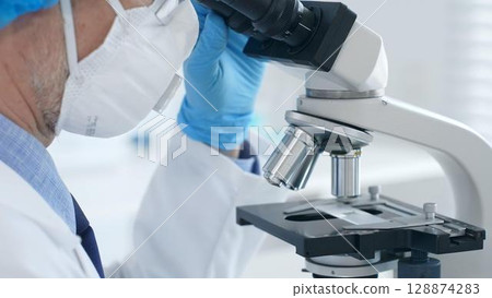 Senior male scientist wearing protective mask, cap and gloves using microscope in laboratory, conducting medical research and analysis. Medicine, healthcare and science concept 128874283