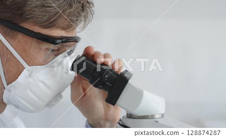A researcher in protective gear examines a sample under a microscope, conducting a detailed study in a sterile laboratory environment. Concept of medicine and science 128874287