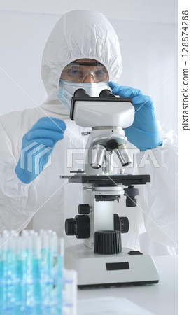 Medical scientist in protective suit using microscope while analyzing samples, conducting research in sterile laboratory environment. Close-up of test tubes with blue liquid Medical scientist in protective suit using microscope while analyzing samples, conducting research in sterile laboratory environment. Close-up of test tubes with blue liquid 128874288