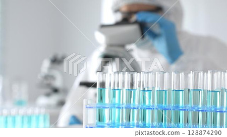 Male medical scientist in protective suit using microscope while analyzing samples, conducting research in laboratory. Close-up of test tubes with blue liquid Male medical scientist in protective suit using microscope while analyzing samples, conducting research in laboratory. Close-up of test tubes with blue liquid 128874290