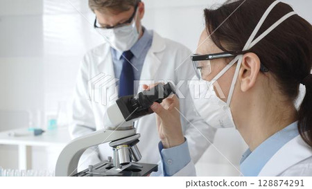 Two scientists, a man and a woman, in lab coats and protective gear, conduct research in a sterile laboratory. Concept of medicine and science 128874291