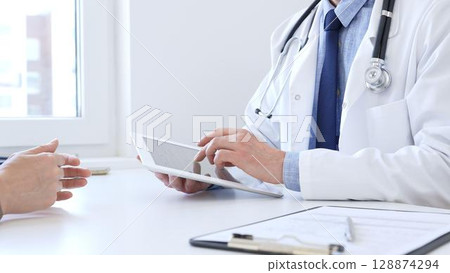 Close-up of doctor man showing information on tablet to patient woman during medical visit, explaining diagnosis and treatment options, providing medical consultation. Medicine concept Close-up of doctor man showing information on tablet to patient woman during medical visit, explaining diagnosis and treatment options, providing medical consultation. Medicine concept 128874294