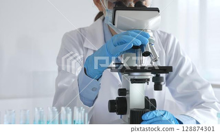 A female scientist in white protective gear adjusts a microscope lens and analyzes medical research samples in a sterile laboratory setting A female scientist in white protective gear adjusts a microscope lens and analyzes medical research samples in a sterile laboratory setting 128874301