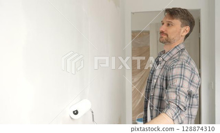 Middle aged man wearing checkered shirt, is rolling white paint on apartment wall with paint roller, transforming interior space with steady strokes during comprehensive home renovation Middle aged man wearing checkered shirt, is rolling white paint on apartment wall with paint roller, transforming interior space with steady strokes during comprehensive home renovation 128874310